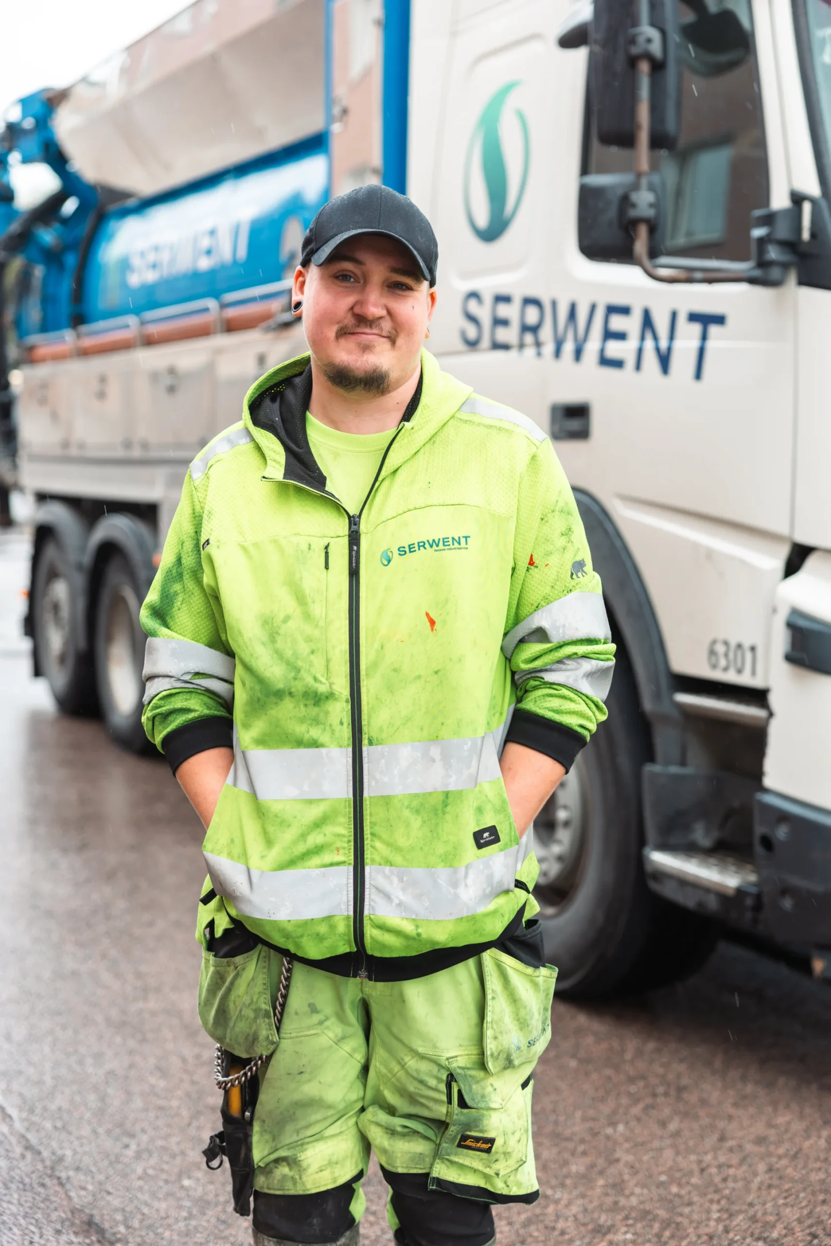 En arbetare i neongrön, reflekterande arbetsuniform står framför en Serwent-lastbil på en våt väg, ler med händerna i fickorna och bär en svart keps.