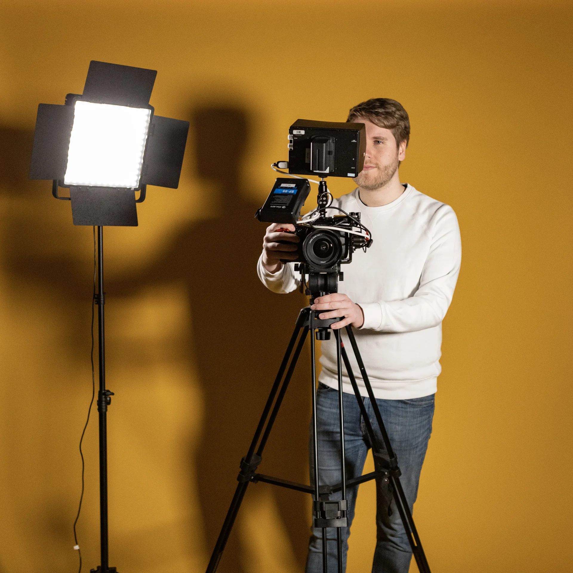 En man i vit tröja manövrerar en videokamera på ett stativ bredvid en stark studiolampa, stående framför en vanlig gul bakgrund.