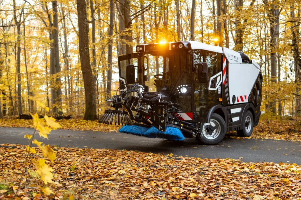Ett sopfordon med blå borstar sopar en asfalterad väg genom en skog med höstlöv på marken och i träden. Starka lampor på fordonets ovansida lyser upp.