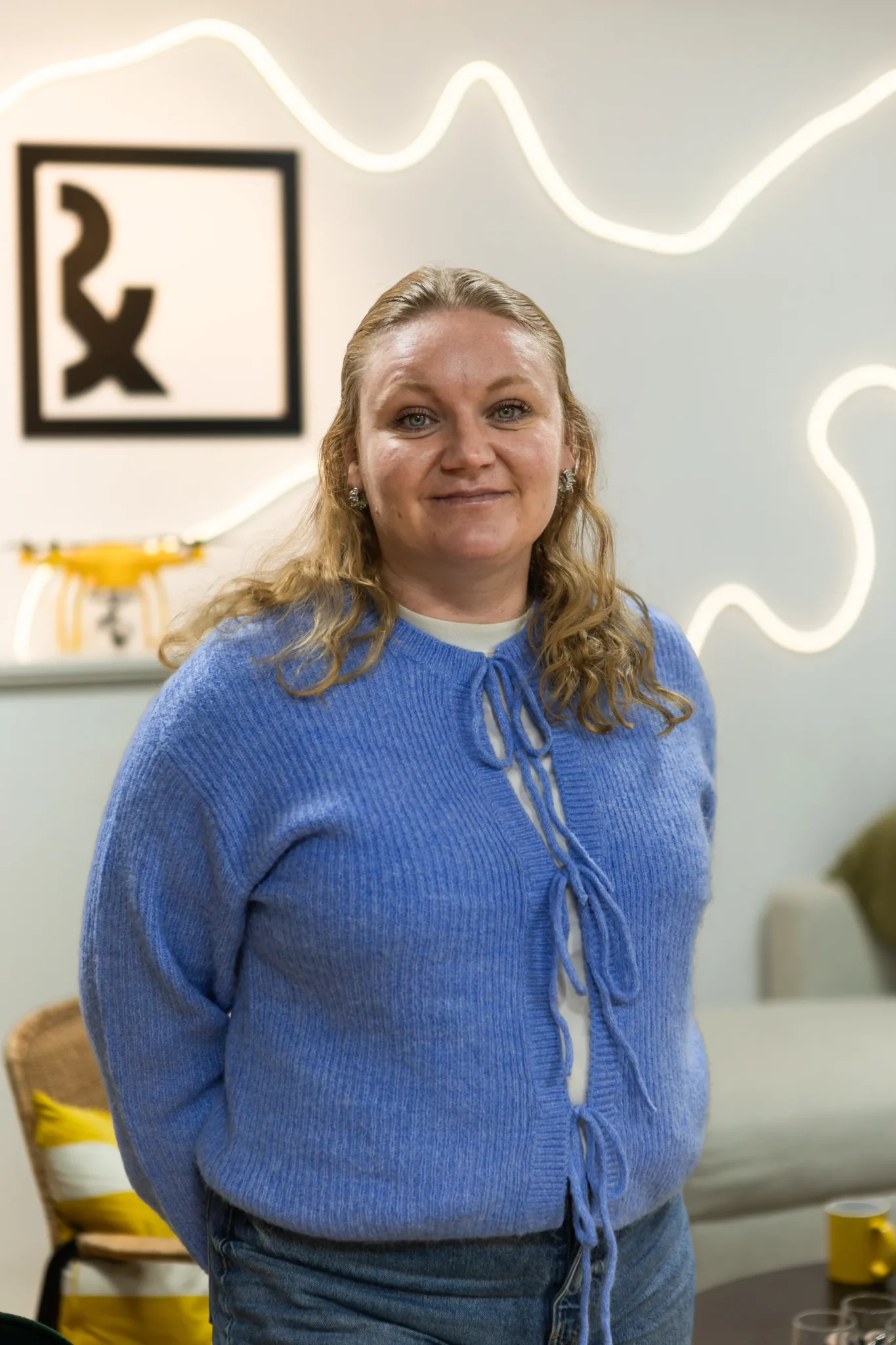 En kvinna med blont hår, ljusblå kofta och vit skjorta står inomhus och ler. Bakom henne syns en neonlampa på väggen och en inramad Rx-symbol. I rummet finns en soffa och gula inredningsdetaljer.