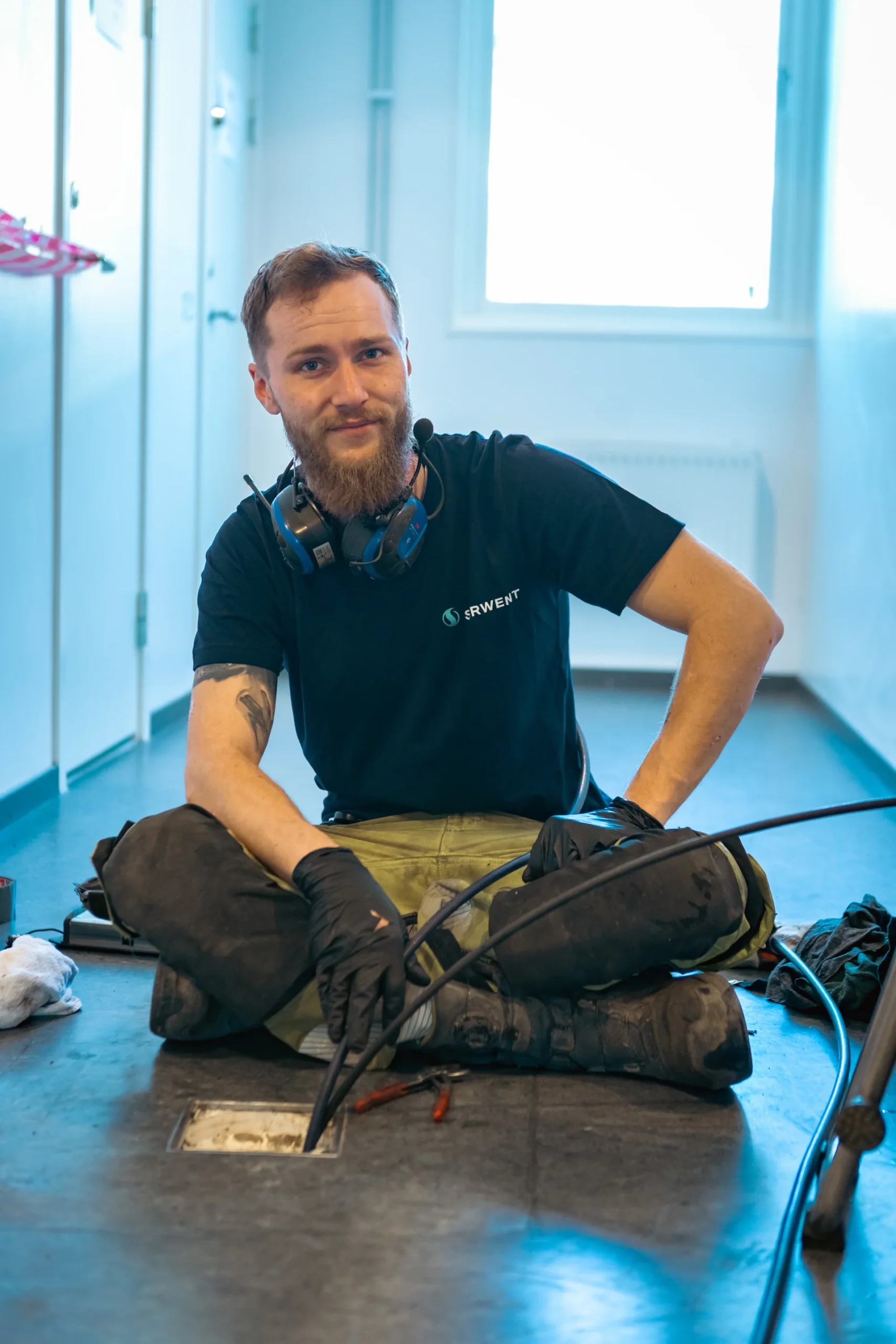 En man med skägg sitter med benen i kors på golvet och bär arbetshandskar, knäskydd, hörlurar runt halsen och en svart t-shirt. Han arbetar med verktyg och kablar i närheten av en öppen golvpanel.
