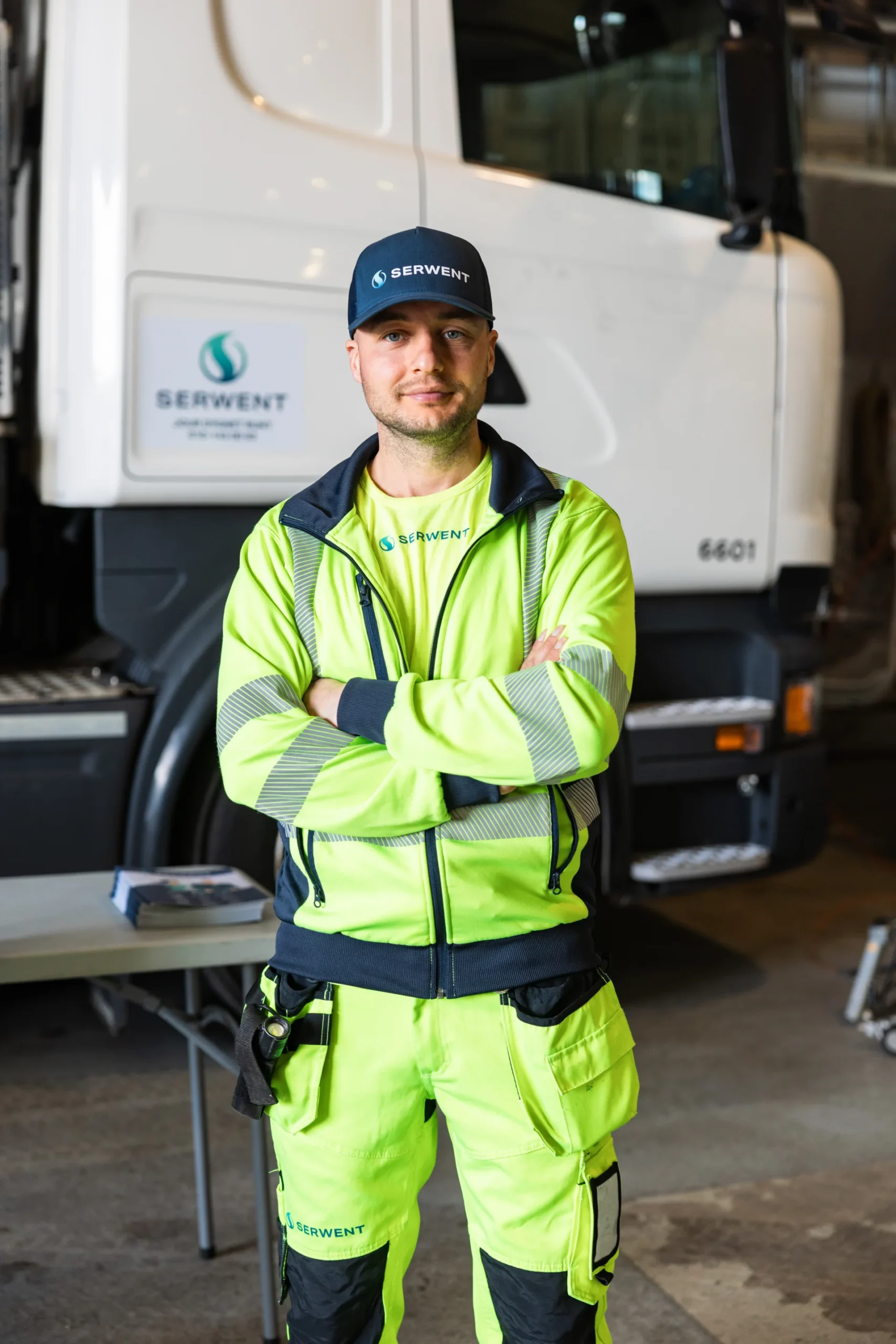 En man i ljusgul arbetsuniform och keps står med armarna i kors framför en vit arbetsbil inne i ett garage. På uniformen och lastbilen syns Serwents logotyp.
