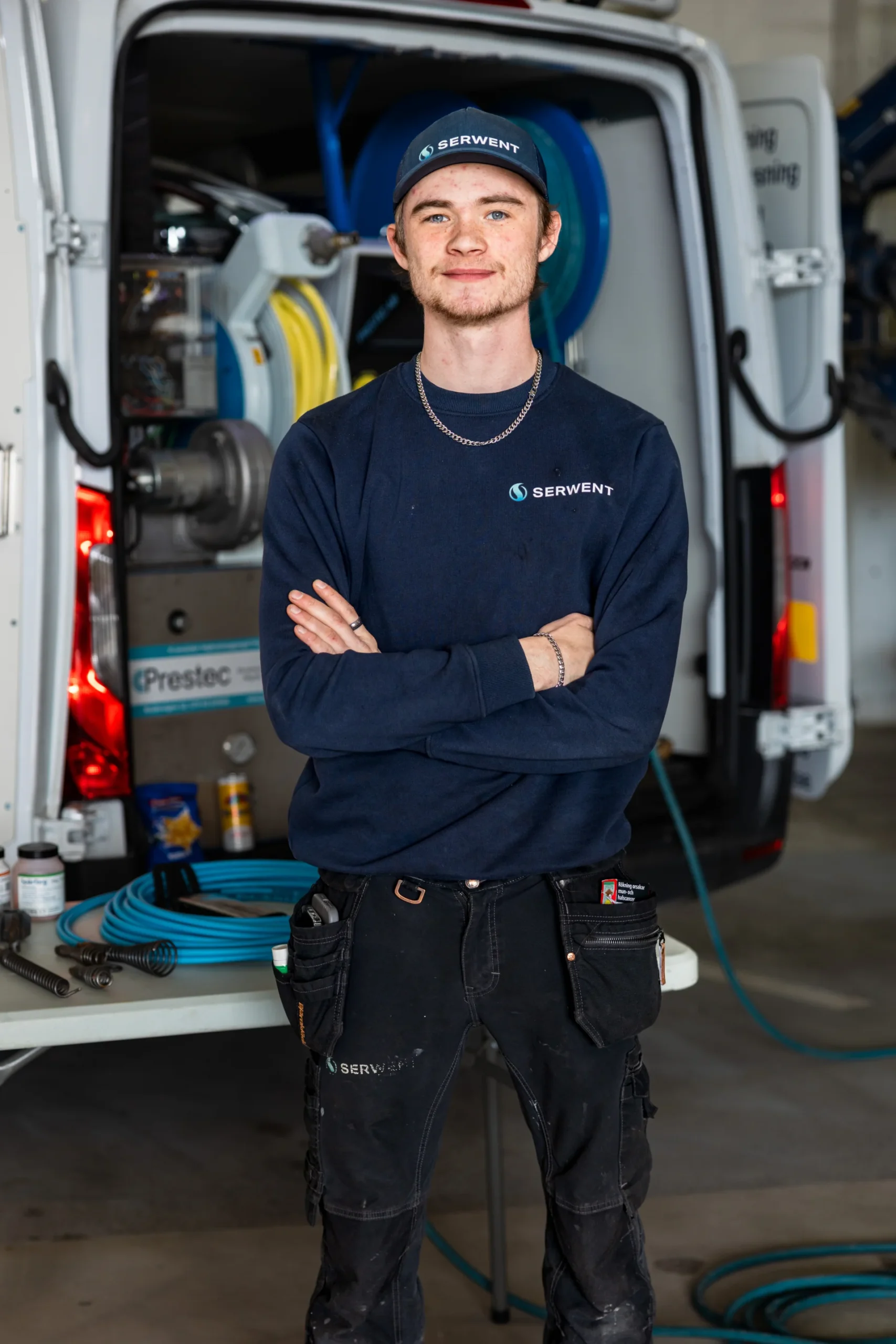 En ung man i arbetskläder står med armarna i kors framför en servicebil fylld med rengöringsutrustning. Han bär en marinblå keps och tröja med Serwent-märkning och ler lite mot kameran.