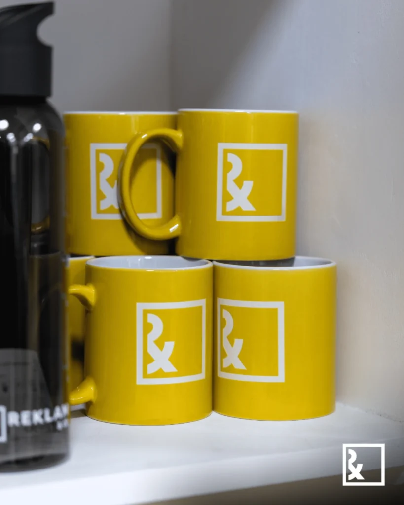 Fyra ljusgula muggar med vita fyrkantiga Rx-logotyper står prydligt staplade på en vit hylla bredvid en svart vattenflaska - perfekt för din nästa livesändning. Muggarna ger en livlig touch till den organiserade hörndisplayen.