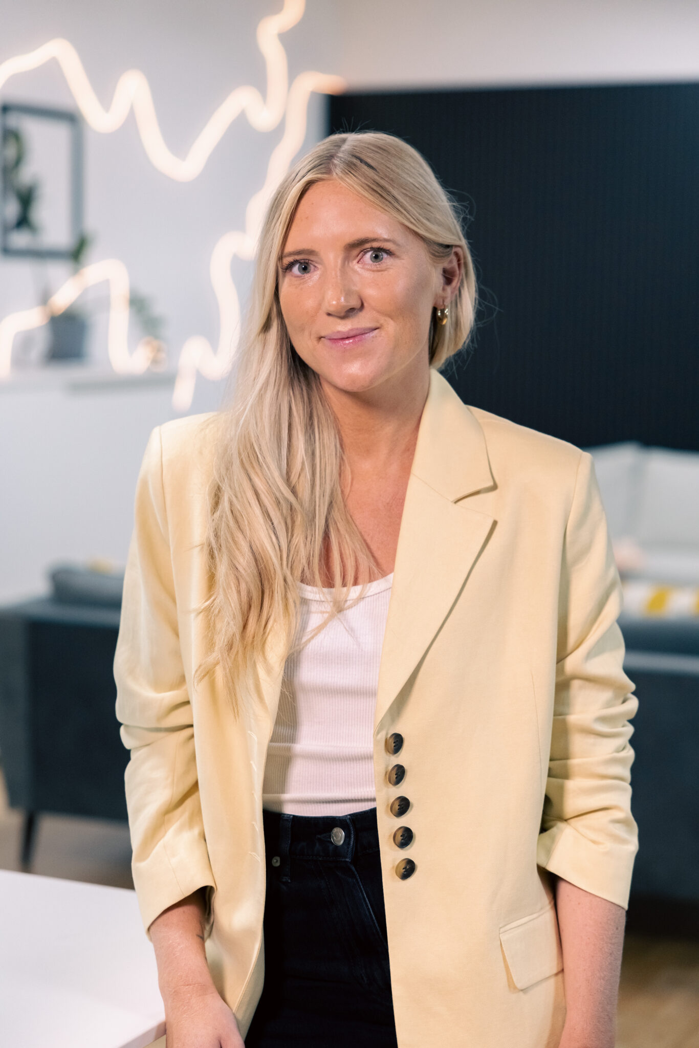 En kvinna med långt blont hår, klädd i en ljusgul kavaj, vit topp och svarta byxor står inomhus och ler mjukt. Bakgrunden har modern inredning med abstrakt belysning och en soffa.