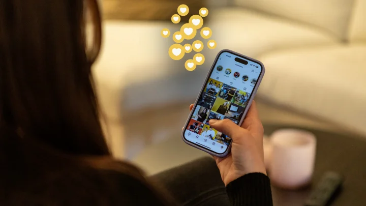 En person som håller i en smartphone och bläddrar igenom ett flöde på sociala medier, med flytande hjärtsymboler ovanför telefonen, sitter i en soffa med ett stearinljus och TV-fjärrkontrollen i närheten.