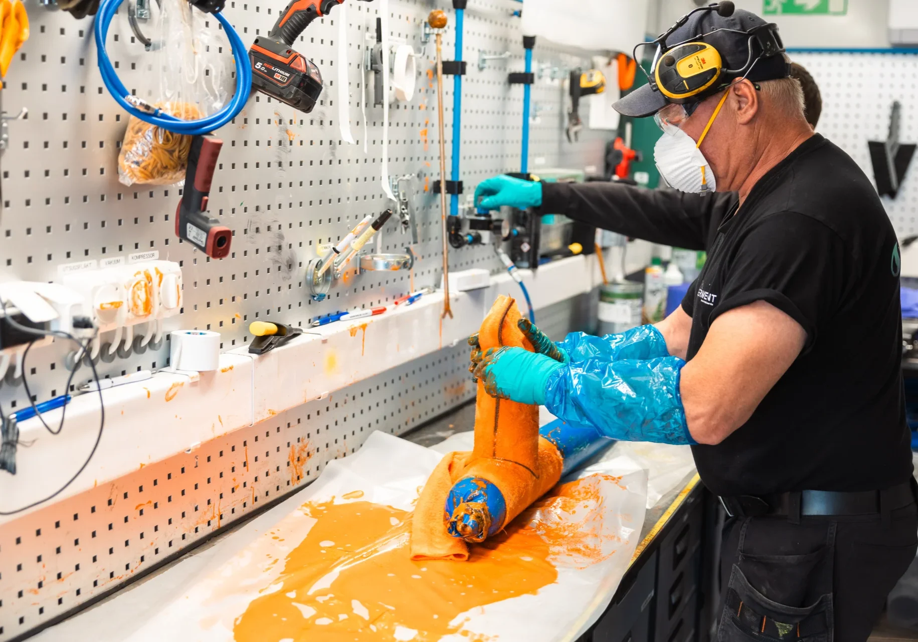 En person iförd handskar, mask och hörselskydd arbetar vid en bänk och hanterar ett orangefärgat föremål med verktyg och utrustning på väggen i en verkstad. Orange vätska stänker ut på arbetsytan.