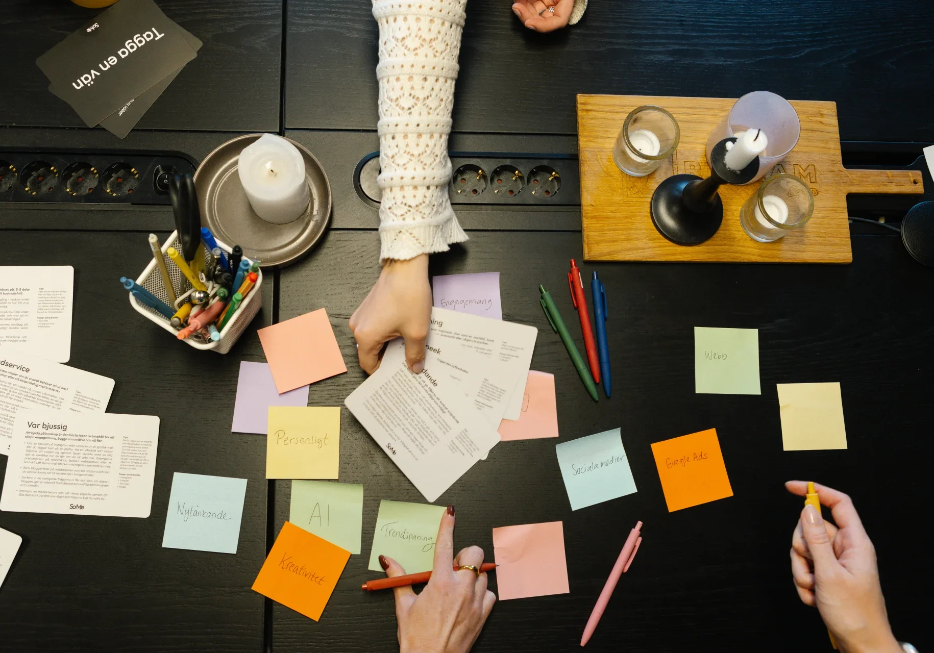 Tre personer arbetar vid ett bord som är täckt med post-it-lappar, papper, pennor och ljus. En person pekar på ett dokument, medan de andra skriver på lappar och håller i pennor, vilket tyder på en gemensam planeringssession.