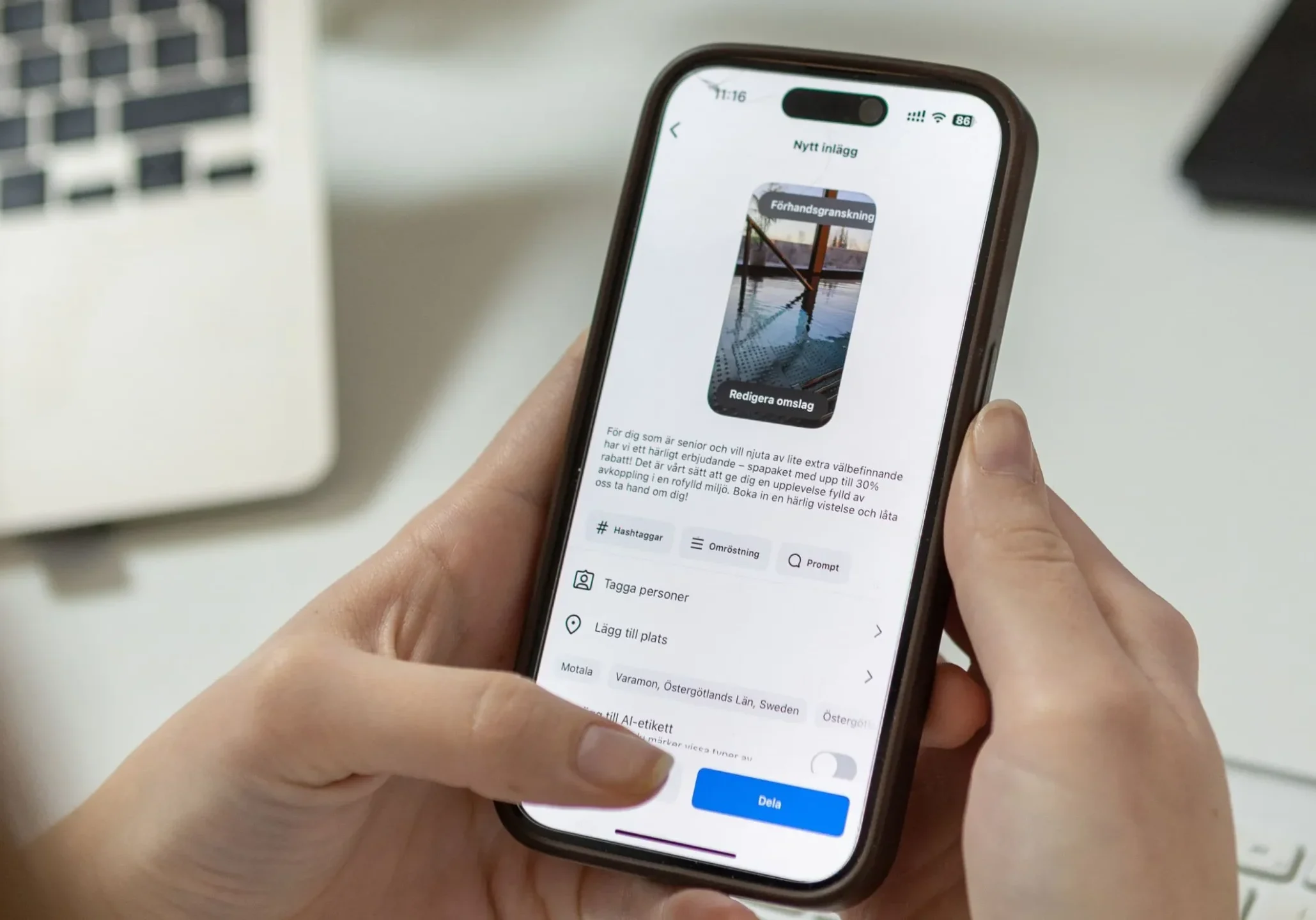 En person håller i en smartphone och redigerar ett inlägg på sociala medier med ett foto av en pir över vatten. En bärbar dator och ett tangentbord syns i den suddiga bakgrunden.
