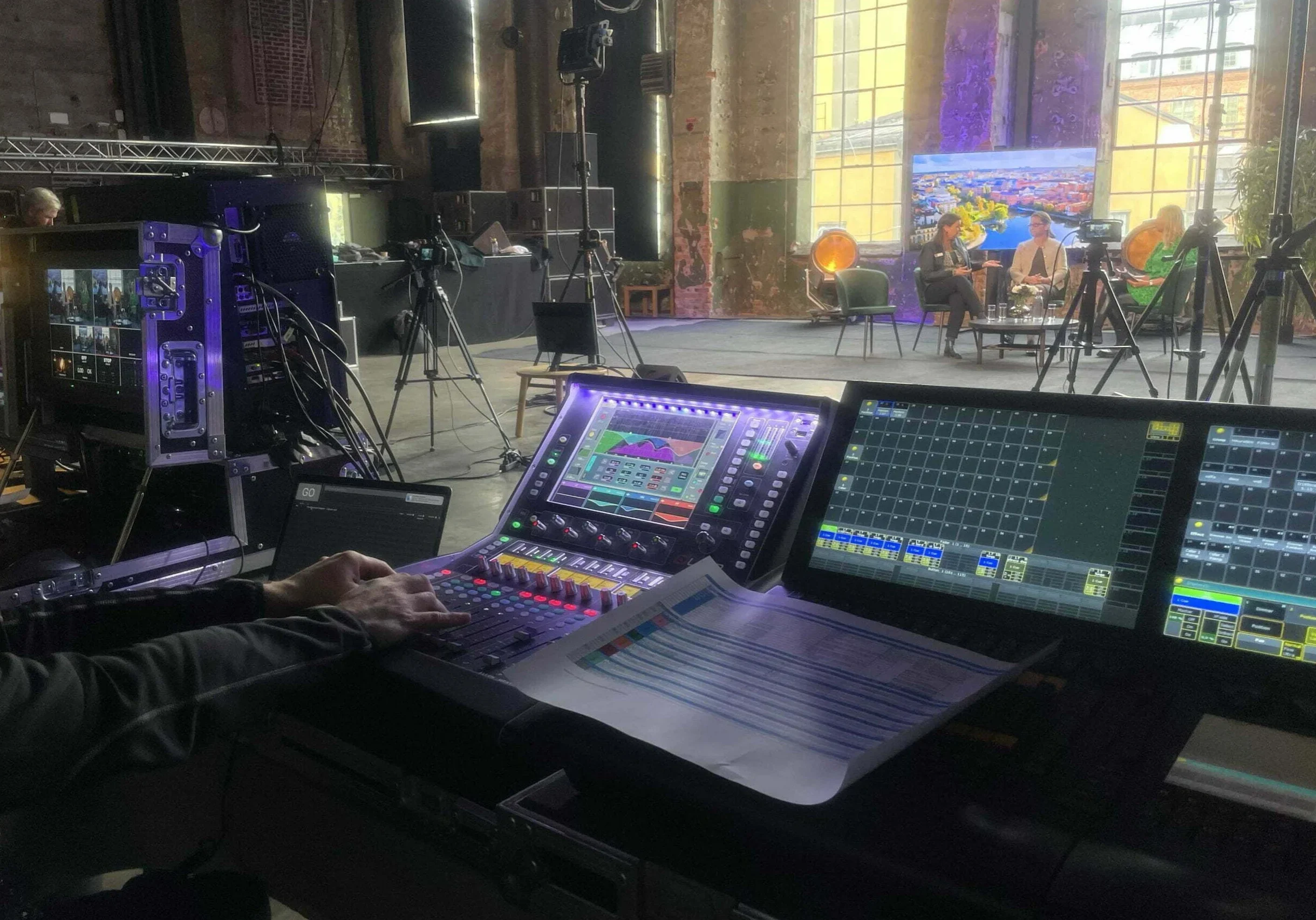 En person sköter en ljudmixerkonsol i en studio med kameror, monitorer och belysning. I bakgrunden sitter människor på en scen framför stora fönster och en färgglad väggmålning.